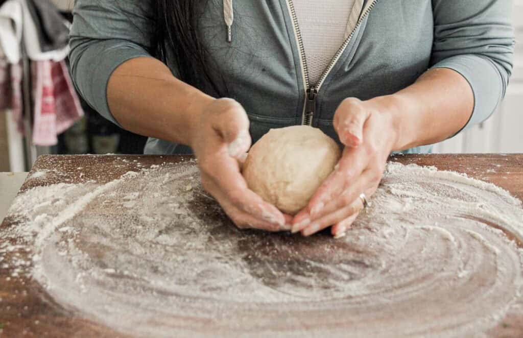 Person shaping a ball of dough with their hands on a floured wooden surface. They are wearing a grey zip-up hoodie, and the kitchen background is slightly blurred.